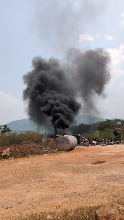 The image depicts a thick plume of black smoke rising from a cylindrical structure and exhaust pipe on a dirt road near Suối Thông A1, Vietnam. Debris and construction materials are visible in the background. A mountain range is faintly visible in the distance. The sky is partly cloudy.