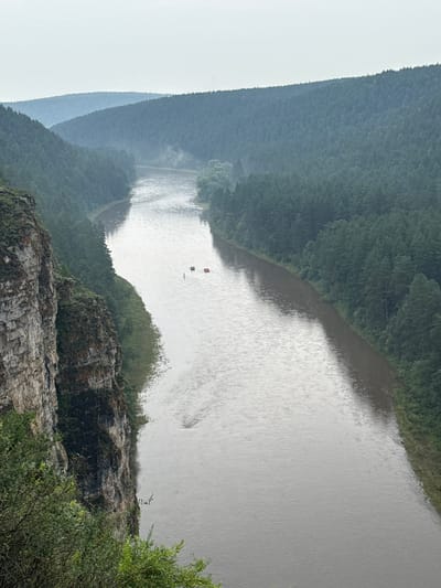This is a high-angle, medium shot of a wide, muddy river flowing through a forested valley on an overcast day. A steep, rocky cliff face with some sparse vegetation lines the left side of the frame, dropping down to the river's edge. The right side of the river is bordered by a dense forest of dark green coniferous trees that stretch up the hillsides. In the distance, the hills recede and become shrouded in mist, with multiple layers of blue-hued, forested mountains visible. On the river, approximately mid-frame, are three small, orange-red inflatable rafts or boats, each with at least one person visible, suggesting recreational activity. The water is choppy with ripples and some surface debris is visible. The overall atmosphere is cool and damp. The location is identified as the city of Kul'metovo, Russia.