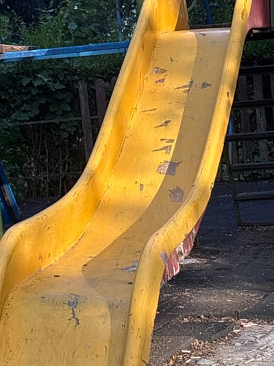 The image shows a weathered yellow plastic slide on an outdoor playground. The slide is curved and has patches where the yellow surface is worn away, revealing a reddish-brown underlayer in some areas. The ground beneath the slide appears to be a dark, cracked surface, possibly asphalt, littered with small pieces of debris. In the background, lush green foliage and trees are visible, suggesting a natural environment. To the right of the slide, a dark, wooden ladder with horizontal rungs leads up to the playground structure. The scene is illuminated by sunlight, casting shadows and highlighting the texture of the slide. This playground is located in the village of Garmen, Bulgaria.