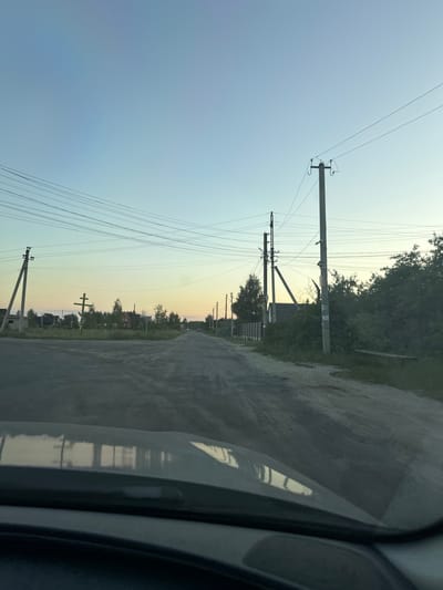 The image depicts a rural road in Orlivka, Ukraine, likely during the late afternoon or early evening, given the soft, gradient sky. The road, appearing to be unpaved or gravel, stretches into the distance, flanked by trees and vegetation. Electrical poles with numerous wires are a prominent feature, running along the side of the road and disappearing into the horizon. In the background, houses and other structures suggest a small village or settlement. A distinct feature on the left side of the road is a large, ornate Orthodox Christian cross. The perspective of the shot is from inside a vehicle, with the side mirror and part of the car's interior visible on the right edge of the frame. The overall mood is serene and quiet, characteristic of a peaceful rural landscape at dusk. There is no visible text in the image.