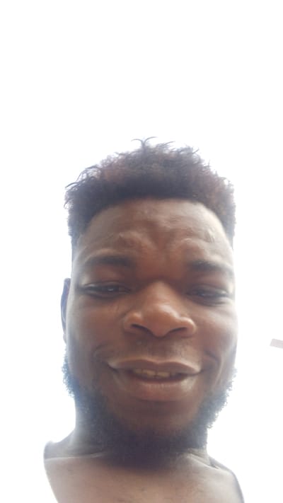 A close-up, low-angle portrait depicts a man with dark skin, short dark curly hair, and a trimmed beard. He is looking directly at the camera with open eyes and a faint, closed-lip smile, revealing his upper teeth. Visible wrinkles are present on his forehead, likely due to the camera angle or his expression. He appears to be shirtless, with his upper chest visible at the bottom of the frame.
The background is entirely overexposed, appearing as a bright white, suggesting an outdoor setting under strong overhead daylight, such as a clear sky. This backlighting causes the man's face to be slightly underexposed, but his features remain clear. The image conveys a casual, self-taken photograph. No specific objects or text are visible in the scene. The context indicates the photo was taken in Enugwu-Ukwu, Nigeria.