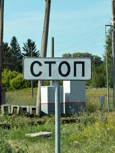 The image shows a stop sign in Russian, which reads "СТОП," mounted on a pole. The sign is rectangular with rounded corners and has black Cyrillic letters on a white reflective background. The sign is positioned in a rural or semi-rural environment, with grassy fields, trees, and power lines visible. In the background, there are utility boxes, concrete structures, and railroad tracks. The weather appears to be clear and sunny. The location is identified as Bezmenovo, Russia.