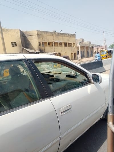 The scene is a street in Chiranchi, Nigeria, under a clear, bright sky. In the immediate foreground, the right side of a white four-door sedan is visible, showing the front and rear passenger doors. The vehicle's interior is partially visible through the front passenger window, revealing a dark headrest on the driver's seat and items on the dashboard. A chrome door handle is present on the front passenger door. Beyond the sedan, a concrete barrier separates the foreground from the main roadway. On the road, a person in blue attire is seated on a light-colored scooter, positioned near the barrier. Behind them, a yellow three-wheeled auto-rickshaw is partially visible. Further down the street, an individual dressed in light-colored traditional clothing walks away from the viewer. The background is dominated by a two-story, light-colored building with multiple rectangular window openings and roof-mounted streetlights. Additional smaller, single-story structures line the street. Multiple overhead utility lines traverse the sky. The buildings appear weathered, indicating a functional urban environment on a dry, sunny day.