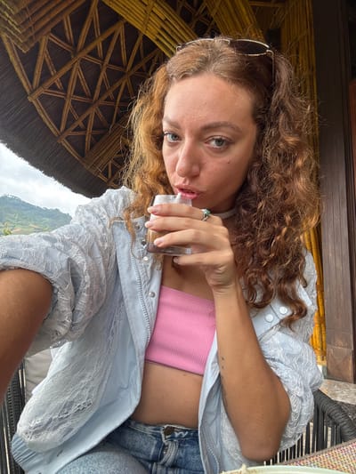 A young adult female is depicted in a close-up, selfie-style shot, seated in an open-air, sheltered structure in Púng Luông, Vietnam. She has shoulder-length, reddish-brown, wavy hair and light green or hazel eyes, directly engaging the camera. Sunglasses with dark lenses and a metallic frame are pushed up onto her head. She is wearing a light blue bomber-style jacket with a distinctive white lace overlay on the sleeves and front panels, zipped up partway over a ribbed, bright pink crop top. Blue denim jeans are visible below the jacket. A silver-toned ring with a prominent light green or turquoise gemstone is on her right ring finger. She is captured mid-action, holding a clear glass containing a dark reddish-brown liquid with her right hand and sipping from it through a transparent straw.
The environment features a prominent thatched roof overhead, supported by a distinct pattern of dark golden-brown woven bamboo lattice. To the left, a lush, verdant hilly landscape under an overcast sky extends into the distance, typical of the mountainous region of Púng Luông. To the right, a dark wooden wall or door frame is visible, suggesting an adjacent building structure, with more bamboo elements. The woman is seated in a dark, woven chair, and a portion of a multicolored, woven tablecloth is visible on the bottom right.