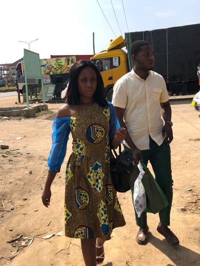 Two adult individuals, one female and one male, are walking on a dry, unpaved ground adjacent to a road in Gbawe, Ghana, under clear daylight conditions.

The female, positioned on the left, has dark hair and wears a brown, yellow, and blue patterned dress featuring an asymmetrical neckline—exposed left shoulder with a sheer blue long sleeve on the right. She carries a black handbag on her left shoulder. Her gaze is directed forward.

The male, positioned to her right, has short dark hair and a beard. He wears a short-sleeved, collared light yellow shirt and dark green trousers. He holds a white patterned plastic bag and a green fabric bag in his left hand. His gaze is directed towards the right side of the frame.

In the background, a large yellow truck with its cab tilted forward is parked. Its cargo bed is covered with a black tarp. To the left of the yellow truck, a roadside stall with a large banner displaying images of fruits (bananas, pineapples, oranges) and contact information, including "0557 0** ***," is visible. A light green signboard frame stands in front of the stall. A paved road is visible to the left, with a partially obscured white truck further in the distance. A portion of a white vehicle is visible on the far right. The ground is light brown earth with scattered debris. Strong sunlight casts distinct shadows to the right of the subjects.