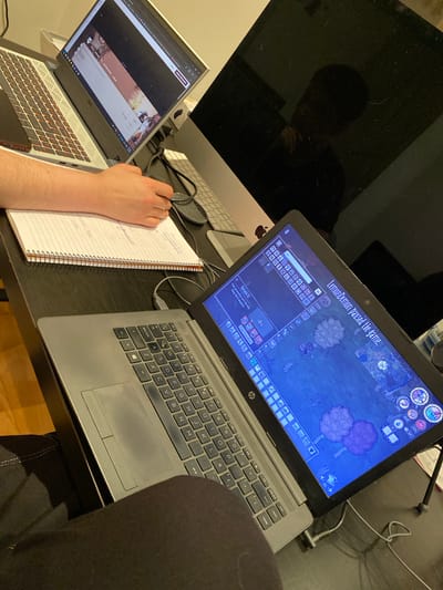The image captures an overhead view of a multi-screen computer setup in an indoor setting, likely a home office or gaming station, located in Villaviciosa, Spain.
The primary subject is a person, whose right arm and hand are visible, resting on a spiral-bound notebook and actively writing. The rest of the person is largely obscured by the camera's perspective and a dark chair/clothing in the foreground, though a faint reflection of their head and shoulders is visible on the large, dark monitor to the left.
The desk holds three computer displays: a large, dark-screened Apple iMac monitor on the left, a silver Dell laptop to its right, and a grey HP laptop further to the right. The Dell laptop is open, displaying what appears to be a dark-themed streaming platform or social media page with some content partially visible. Underneath the Dell laptop, a smartphone is partially visible. The HP laptop is open and prominently displays a video game, "Don't Starve Together," which is currently paused. A message "LemonDemon paused the game." is clearly visible, along with game UI elements showing character stats (health 106, hunger 200, sanity 135) and the season as "Autumn." A crafting menu with items like a "Nautopilot Kit" and a description "Your personality isn't the only thing that's magnetic." is also open.
Notable details include the clutter of cables behind the laptops, connecting them to power or peripherals. The HP laptop's keyboard has a QWERTY layout with keys like "repág" and "imp pant" visible. The Dell laptop features a backlit keyboard. The notebook contains handwritten notes, including numbers like "x 450," "7200," and "4950." The overall lighting suggests an artificial indoor environment, potentially evening or night.