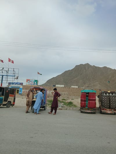 This image depicts a street scene in what is likely the city of Bostan, Pakistan. Three men are gathered near a row of decorated auto-rickshaws ("tuk-tuks"). One man, dressed in a maroon robe, is walking toward the rickshaws. The second man is dressed in blue, and the third man is in a tan-colored suit. The background includes a tan mountain, a brick wall with posters, and Pakistani flags. Overhead utility lines are visible. The road appears to be paved.