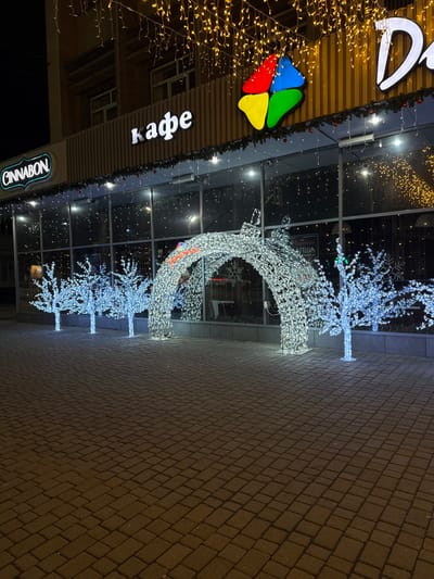 A sideways view showcases a building façade with glass windows and doors adorned with Christmas lights and decorations. Notably, a white, illuminated reindeer and tree display stands on the sidewalk. The building features signage for a café and the brand "Cinnabon." A multi-colored logo is also present. The sidewalk is constructed of brick pavers.