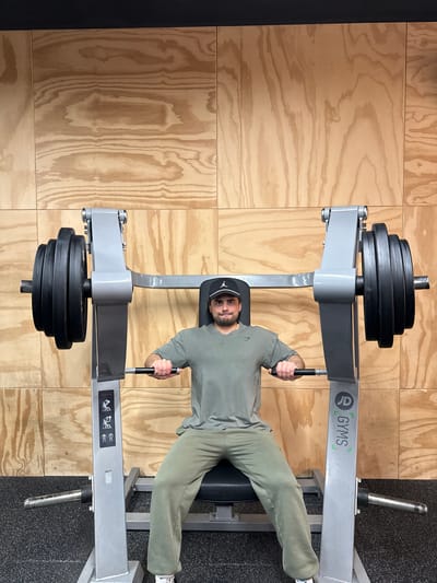 An adult male is seated on a grey plate-loaded chest press machine in Gyms Coventry, United Kingdom. The man, with short dark hair, a beard, and a mustache, wears a black baseball cap with a white Jordan logo, an olive-green t-shirt, and olive-green sweatpants. He grips the machine's handles. Each side of the machine is loaded with five black circular weight plates. The machine bears a "Chest Press" instructional label and a vertical "G GYMS Coventry" logo. The man looks directly forward. The background is a wall of light-colored plywood panels, with a black strip running along the top. The floor is dark grey, textured rubber gym flooring.