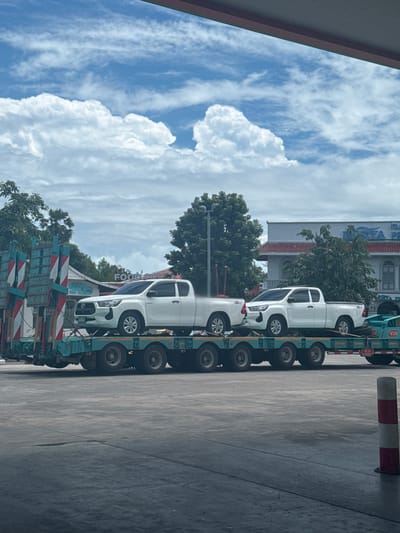 The image depicts a truck carrying several white pickup trucks on a flatbed trailer. The truck is parked on a paved surface, possibly a road or a parking lot. The background shows buildings, trees, and a cloudy blue sky. The time of day appears to be daytime, and the weather is likely overcast or partly cloudy. Some text is visible on the buildings, but it is not clearly legible. The overall scene suggests a transport or delivery operation. The context provided indicates the location is Ban Yid, Thailand.