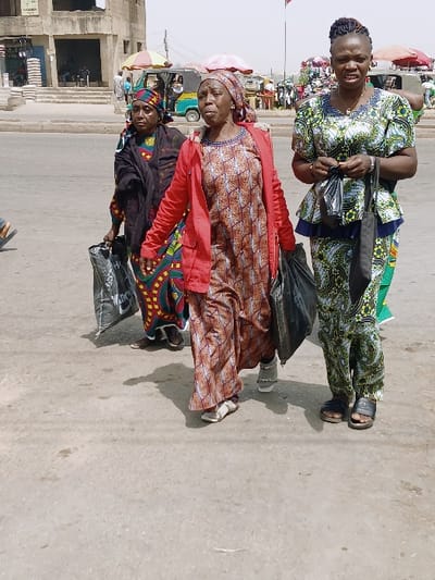 Three adult women are depicted walking on a concrete street in Masvingo, Zimbabwe. The woman on the left wears a multi-colored patterned headwrap, a dark long-sleeved top, and a vibrant patterned long skirt or dress. She carries a large black plastic bag in her left hand. The central woman wears a patterned headwrap, a bright red long-sleeved jacket over a brown and white patterned long dress, and carries a large black plastic bag in her left hand. Her mouth is slightly open. The woman on the right has braided hair and wears a green, black, and white patterned two-piece outfit. She carries a small black plastic bag in her left hand. All three women are looking forward and appear to be walking towards the viewer's right.

In the background, a multi-story, light-colored concrete building with open ground-floor areas is visible on the left. Further back, a light green and yellow auto-rickshaw (tuk-tuk) is present. Colorful market umbrellas (red and yellow) indicate market stalls, and other individuals are visible in the distance, suggesting a busy street or market environment. The ground is light grey concrete. The sky is clear, indicating daylight.
