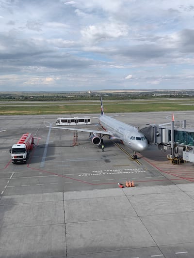 This aerial view captures an airport tarmac on a cloudy day. The primary subject is an Aeroflot airplane docked at a jet bridge. The plane is silver with the Aeroflot logo and text visible on its side. A jet bridge is connected to the aircraft, and there appears to be at least one person near the plane's entrance.
In the foreground, a red and white fuel truck is parked next to the airplane, connected by a hose, indicating that refueling is in progress. Further back on the tarmac, a white airport bus with tinted windows is visible. The scene is set on a concrete tarmac with painted lines and markings, including what appears to be a "CUSTOMS CONTROL ZONE" sign in Russian.
The background shows a flat landscape with green fields and some distant buildings, suggesting a rural or semi-urban airport setting. The sky is filled with a mix of gray and white clouds, indicating overcast conditions. The overall impression is one of an active but perhaps slow-paced airport operation. The time of day is difficult to pinpoint precisely due to the cloud cover, but it appears to be daytime. There are no obvious emotional cues conveyed, and the scene is factual and functional.
