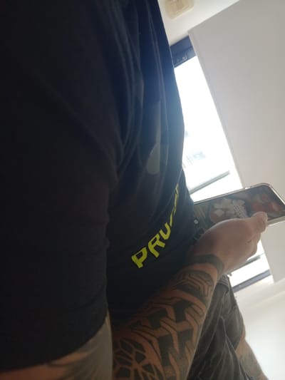 A person is lying down, holding a smartphone in their hand. The smartphone is displaying a video, possibly from TikTok, showing food being prepared. The person has tattoos on their forearm, including a large geometric design. They are wearing a dark-colored garment with the word "PRU" visible in yellow lettering. The setting is indoors, next to a window with daylight coming through. The location is identified as Lomas de Zamora, Argentina.