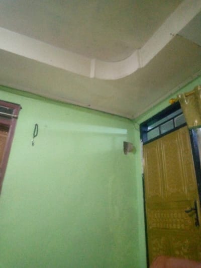 The image depicts the interior corner of a room, located in Kuningan, Indonesia. The walls are painted a uniform light green color. The ceiling is off-white and features a white decorative plaster molding that follows the wall-ceiling joint and curves around the corner. On the left wall, a partial view of a brown-framed window or opening is visible. A black hook or loop is affixed to the left wall, from which a single key is suspended. Towards the right wall, a small, light-brown, triangular bracket is mounted. On the far right, a portion of a dark-framed door is visible, featuring a lighter, possibly gold-toned, panel with embossed vertical and horizontal patterns. A black door handle is present on this door. Above the main door panel, a transom window with multiple panes is integrated into the door frame. No individuals are present in the scene.