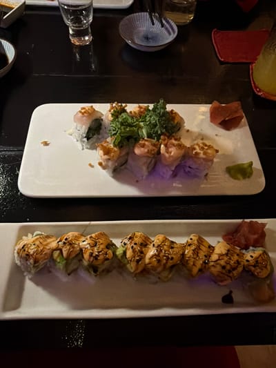 The image shows two plates of sushi on a dark, reflective surface, likely a table. The top plate contains several pieces of sushi rolls, some topped with a creamy sauce and crispy bits, with a garnish of green leaves in the center. There is also a small pile of pink pickled ginger and a dollop of green wasabi on this plate. The bottom plate is filled with a line of sushi rolls, also topped with a creamy sauce and sesame seeds. This plate also has pickled ginger on the right side. In the background, a small shot glass and a blue and white patterned bowl are visible. To the right, a red cloth and a glass with a yellow beverage are present. This scene is consistent with a dining establishment, potentially a restaurant or bar in San Pedro Town, Belize, given the common presence of sushi restaurants in tourist destinations. No people are visible in the image.