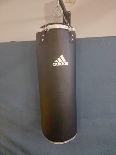 A black Adidas punching bag hangs from a metal bracket mounted on a light-colored wall. The punching bag is cylindrical and has white trim at the top and bottom. The Adidas logo is visible in white on the front of the bag. The background consists of the wall and a dark blue surface that appears to be a padded wall or mat. The image is taken from a medium distance. There are no people or animals present in the scene. The lighting is dim and appears to be artificial. This scene is not located in Nuremberg, Germany.