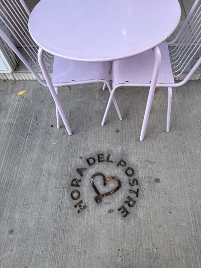 The scene captures a section of a gray concrete sidewalk outside a business. The foreground is dominated by a round lavender-colored metal table and chairs. To the right, text is embedded in the concrete, spelled out using dark metallic or resin letters, spelling "DEL POSTRE HORA" in a curved arrangement, with a stylized heart in the center. This is related to "La Hora del Postre" located in the city of Salta, Argentina. The environment seems to be an outdoor seating area of a café or restaurant.