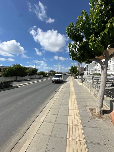 The image depicts a street scene in Paphos Municipality, Cyprus, on a bright, sunny day with a blue sky dotted with white cumulus clouds. The perspective is from a sidewalk looking down a street lined with trees and buildings.
On the right side of the image, a sidewalk is visible, paved with grey and yellow tactile tiles. A tree with a thick, light-colored trunk and lush green foliage stands prominently on the sidewalk. Beyond the tree, there's a metal railing and outdoor seating areas with tables and benches, suggesting a commercial or dining establishment. White buildings with multiple levels are situated behind the railing.
The street itself is paved with asphalt and shows some wear. A white van is driving on the left side of the road. Further down the street, other vehicles and pedestrians can be seen. Trees and white fencing line the opposite side of the street, with buildings in the background. Streetlights are also visible along the roadside. The environment appears to be an urban or suburban area, possibly a commercial district.