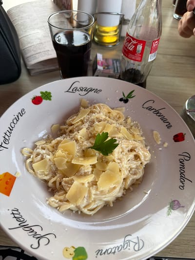 The image shows a plate of pasta, likely fettuccine or spaghetti carbonara, served with a creamy sauce and topped with shaved parmesan cheese and a sprig of parsley. The plate is decorated with various pasta names and illustrations, including "Fettuccine," "Lasagne," "Carbonara," "Pomodoro," and "Spaghetti."

On the table, there is a glass of dark liquid, possibly cola, next to a partially consumed bottle of Coca-Cola. A pack of cigarettes is also visible. In the background, a menu or newspaper is partially obscured by a black handbag.

The setting appears to be an outdoor cafe or restaurant with a wooden table. The overall lighting suggests it is daytime. The location is identified as Svishtov, Bulgaria.