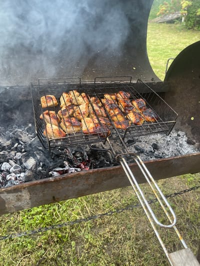 The image shows a close-up, eye-level shot of a barbecue grill filled with charcoal and cooking meat. The grill basket is filled with pieces of meat that appear to be chicken or pork, arranged in two rows. The meat is browned and slightly charred, indicating it is actively being cooked. Smoke is rising from the grill, obscuring the background slightly. The grill itself is made of metal and shows signs of use and rust. The surroundings appear to be an outdoor, grassy area. There is a chain visible in the foreground, lying across the grass, which is slightly blurred. The overall impression is that of an outdoor cooking scene. The location is described as the city of Kalnciems, Latvia, which suggests a rural or semi-rural setting consistent with the grassy environment. There are no people visible in the image.