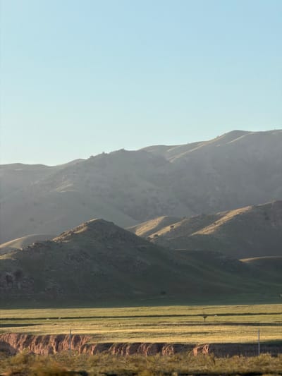 The image shows a landscape in Chüy, Kyrgyzstan. The terrain consists of undulating hills and mountains under a light-blue sky. The right side features a grassy field. A pole structure is visible. The landscape appears dry.