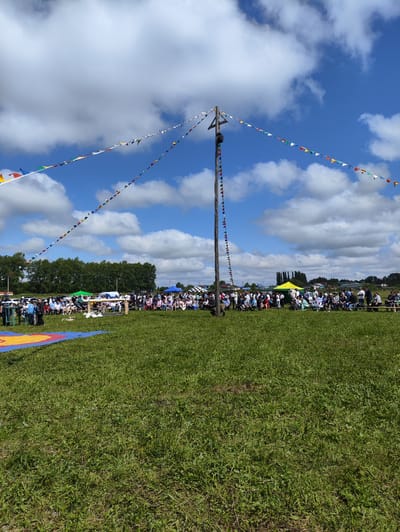 An outdoor event is taking place in a grassy field under a bright, cloudy sky. A tall wooden pole stands prominently in the center, adorned with strings of colorful triangular flags that stretch across the sky. A crowd of people are seated on benches and standing behind a wooden barrier in the middle ground, facing a wrestling mat laid out on the grass in the foreground. Several tents and some vehicles are visible in the background, beyond the crowd. The event appears to be a cultural or sporting gathering, possibly a wrestling competition or festival, taking place in the village of Novo-Ursayevo, Russia.