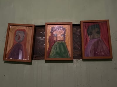 Three framed paintings of abstract figures are displayed side-by-side on a textured, light green wall in Karangkebagusan, Indonesia. The paintings, rendered in what appears to be watercolor or gouache, depict stylized human figures with simplified features. The leftmost painting shows a figure in red and purple with a grey face. The central painting features a figure with dark hair, a red torso, and a green skirt. The rightmost painting depicts a figure with dark hair, a dark face, and a purple garment against a pink background. The frames are made of light brown wood. Behind the paintings, there is a dark, crinkled material, possibly plastic sheeting. The wall shows signs of wear and has some small dark specks.