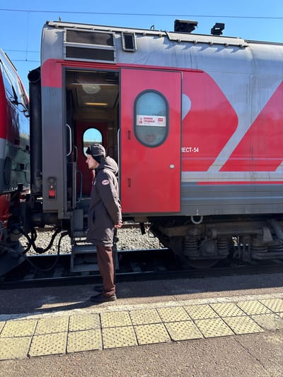 The image displays a stationary railway scene at a train station in Kropachyovo, Russia, under a clear blue sky. A male individual, appearing to be a railway employee, stands on a yellow tactile-tiled platform. He is attired in a dark brown winter coat featuring the Russian Railways (RZD) logo on the left chest, dark trousers, and dark shoes, topped with a dark winter hat. His hands are clasped behind his back, and he is oriented facing left, parallel to the train.
Immediately adjacent to the individual is a red and grey RZD passenger train car. Its main red passenger door, containing a rounded window, is open, revealing the interior vestibule. A sign visible through the window reads "ПОМНИМ ГОРДИМСЯ!" (We remember! We are proud!) accompanied by the RZD logo. The side of the train car features the RZD logo and the text "IECT-54". Another train car, primarily red and dark grey, is partially visible on the left, connected to the described car. Beneath the train car, railway tracks laid on gravel ballast are discernible. The platform transitions from yellow tactile tiles to a darker, solid surface. The environment is illuminated by direct sunlight, producing clear shadows.