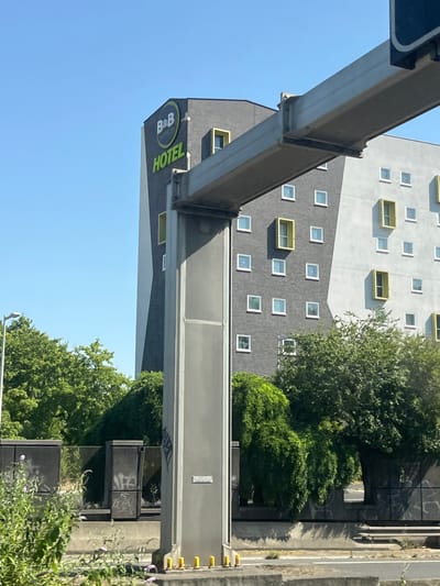 A B&B Hotels building is visible under a clear blue sky in Nanterre, France. The hotel features a dark gray facade with lighter gray and white geometric sections, punctuated by numerous small white square windows and several recessed yellow rectangular windows. A prominent sign on the building reads "B&B HOTELS" in green lettering. In the foreground, a large, gray metal structure, likely part of an overpass or highway signage, extends across the frame. The base of this structure is supported by concrete pillars with yellow accents. Lush green trees partially obscure the lower floors of the hotel and a lower wall with graffiti. A street lamp is visible to the left of the hotel, and a section of a road is present in the bottom portion of the image. The overall scene appears to be a snapshot taken from a vehicle traveling along a road, looking up at the hotel and overpass.