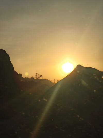 The image depicts a sunset over a quarry in Riba-roja de Túria, Spain. The sun, a bright, intense orb, is positioned slightly off-center to the right, casting a warm golden glow across the sky. Silhouetted against the bright sky are several large, dark mounds of what appears to be excavated material, forming a rugged, mountainous landscape.

In the mid-ground, a substantial industrial structure, likely part of the quarry's operation, is visible. This structure features a prominent conveyor belt system or similar machinery, with tall supports and inclined belts, all rendered in silhouette. Further back, a few scattered trees can be seen, also silhouetted, and a few utility poles with visible wires suggest the presence of nearby infrastructure.

The foreground is dominated by dark, shadowy shapes, likely more piles of excavated material or the edge of the quarry itself. Scattered throughout the image, especially in the foreground and mid-ground, are numerous out-of-focus circular or polygonal shapes, which appear to be lens flares or bokeh effects caused by the light source and potentially dust or imperfections on a lens. A distinct sunbeam or lens flare cuts diagonally across the image from the sun towards the bottom left. The overall atmosphere is one of dramatic natural light interacting with an industrial environment. There are no people or animals visible in the scene.