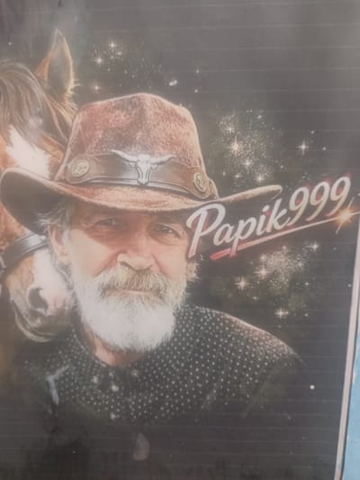 The image is a medium shot of a man wearing a cowboy hat and a polka-dot shirt. He has a white beard and is looking towards the camera. A horse is partially visible behind him on the left side of the image. The background appears to be a starry sky with a gradient of dark blue to black. There is a signature or watermark that reads "Papik999" in cursive script with a red outline, placed over the man's hat and shoulder. The image is a photograph, possibly a portrait or a digital artwork. There is no visible indication of the location being the city of Maloye Lugovoye, Russia.