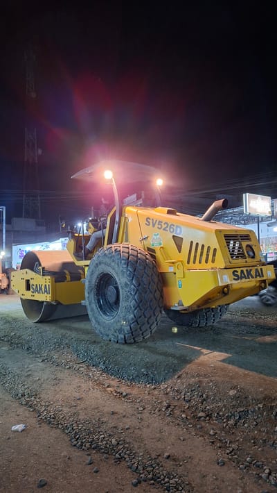 A yellow Sakai SV526D roller is parked on a construction site at night in Blangpeuria, Indonesia. The roller is the primary subject, positioned in the center of the frame, with its large, treaded tire dominating the foreground. A person is seated in the driver's seat of the roller, wearing a jacket and pants. The construction site is characterized by loose gravel and dirt, with evidence of recent work. In the background, the lights of nearby buildings and a cell tower are visible, suggesting an urban or semi-urban environment. The scene is illuminated by the roller's headlights and ambient lighting from the surroundings.