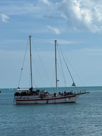 A white and red sailboat with two masts is on calm, blue water under a partly cloudy sky. The boat is carrying passengers who are standing on the deck and appear to be on a tour or excursion. There are flags strung along one of the masts, and a Bulgarian flag is flying from the stern. In the background, two smaller sailboats are visible in the distance. The image appears to have been taken from a pier or another boat. The boat's name, "DNIS," is visible on its side, along with the registration number "BG4 4848." The scene is consistent with the Black Sea coast near Balchik, Bulgaria, which is known for its maritime activities and scenic views.