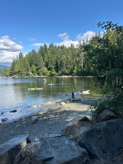 This image depicts a serene lake scene with a backdrop of a dense, green forest and a clear blue sky with scattered clouds. The foreground features a rocky shoreline with large boulders. The lake itself is calm and reflects the sky and surrounding trees.
Several individuals are engaged in water activities on the lake. In the mid-ground, one person is kayaking in a green kayak. Further back, another individual is paddleboarding. There are also at least two other people visible on paddleboards. A small white boat is anchored in the lake near the distant shore. The shoreline on the opposite side of the lake is lined with tall evergreen trees, and several houses are nestled among the trees, suggesting a residential area.
The location is likely in a recreational area within the city of North Vancouver, Canada, known for its natural beauty and proximity to forests and bodies of water. The overall atmosphere is one of peaceful outdoor recreation on a sunny day.
