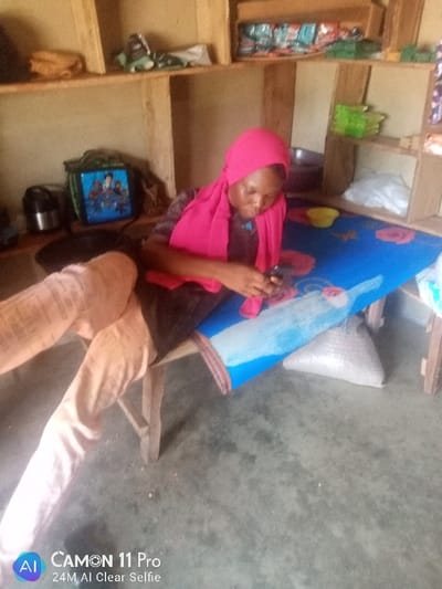 An indoor scene in Unguwan Rimi, Nigeria, shows a female individual seated on a wooden stool, actively engaging with a dark-colored mobile phone held in both hands. She wears a pink headscarf and a dark top, with light-colored trousers on her extended legs. She is positioned beside a low wooden table covered with a blue cloth featuring a floral pattern and a distinct light blue or white painted section. A small yellow bowl rests on the table. In the background, a multi-tiered wooden shelving unit contains various items, including packaged goods, a brown pouch, a green bag displaying a cartoon image, a silver thermos, a green plastic container, and a white plastic bag. A white woven bag is visible on the light gray concrete floor beneath the table. The walls are light-colored, and the environment appears functional.