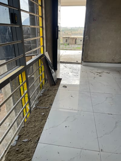 This is a photograph taken at a construction site in Angwan Sayawa, Nigeria. The image is oriented vertically, showing a partially completed building. In the foreground, metal bars form a protective grate over windows, behind which is a glimpse of dark glass. Yellow protective tape with the text "Solidking Aluminium Only for the kings" is visible along the window frames.

Beyond the grate, the scene transitions to interior space. The floor is covered with large, light-colored tiles, some of which are dirty with construction debris. To the left, a concrete wall or ceiling is visible, with some white speckling. An open doorway or entrance leads to an exterior view.

The exterior shows a landscape with hills and scattered buildings under construction. One building in the distance appears to be made of concrete blocks with wooden scaffolding. The sky is overcast and bright, suggesting it is daytime, likely with no specific weather conditions to note. The overall impression is one of ongoing construction and development in a semi-rural or developing urban area.