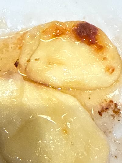 The image shows two cooked pierogi covered in a clear, possibly oily, liquid. The pierogi on the left has a browned or burnt area on its edge. There is some dark residue or crumbs present in the upper right portion of the image. The overall texture appears smooth and slightly glossy due to the liquid coating. There is no visible information in the image that relates to the city of Dania Beach, United States of America.