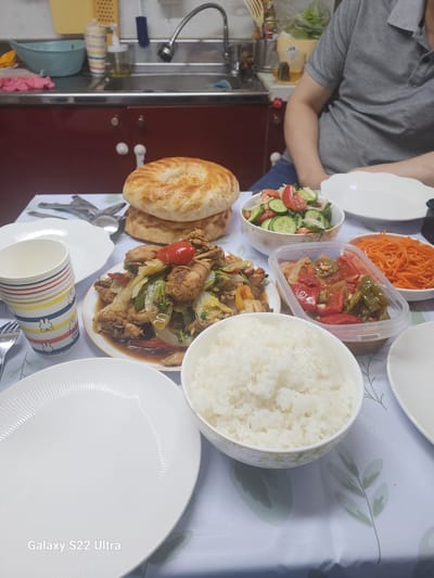 The image shows a table laden with food, likely in a home kitchen or dining area. In the background, a kitchen sink with a faucet is visible, along with some containers and cleaning supplies, suggesting a domestic setting. The table itself is covered with a patterned tablecloth and features several dishes of food. These include a stack of flatbreads, a salad with tomatoes and cucumbers, a dish of stir-fried chicken and vegetables, a container of cooked bell peppers and onions, a bowl of shredded carrots, and a large bowl of white rice. There are also white plates, cutlery, and a paper cup with a colorful pattern. A person wearing a grey t-shirt is partially visible in the upper right corner of the image, suggesting they are seated at the table. The scene depicts a meal being prepared or served. No specific indicators of Ansan, South Korea are present in the image.