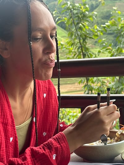 A person is seen in a close-up, three-quarter profile, sitting outdoors and eating in Púng Luông, Vietnam. The individual has dark hair styled in numerous thin braids, some adorned with small silver-colored rings or beads. Their gaze is directed slightly downwards and to the right, with lips subtly pursed as if in the act of chewing or contemplation. They are wearing a textured red garment, possibly a jacket or shirt, featuring small white embroidered hearts. A light green tank top and a thin gold chain necklace are visible underneath the red garment. The person's right hand holds a pair of dark chopsticks with patterned metallic tips, poised over a light gray or beige ceramic bowl filled with food, which appears to be a soup or noodle dish with various ingredients including what look like mushrooms or dumplings. A silver spoon is also present within the bowl. The person's fingernails are long and pointed, with an iridescent sheen. In the blurred background, lush green foliage, trees, and gently sloping hills or mountains are visible, characteristic of a natural, possibly agricultural, landscape. A dark wooden railing crosses the mid-ground, separating the dining area from the expansive view.