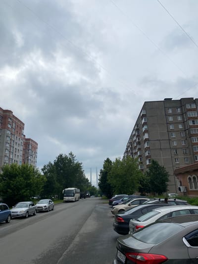 This image captures a street scene in Cherepovets, Russia, under an overcast sky. On the left, a multi-story residential building with a red and grey facade stands tall. Across the street, another large, grey apartment building dominates the right side of the frame, featuring numerous windows. A row of parked cars lines the right side of the street, including a grey sedan and a black sedan in the foreground. Further down the street, a white bus is parked, and beyond that, other vehicles and trees are visible. In the distance, a suspension bridge with tall pylons is faintly visible against the cloudy sky. The street itself appears to be asphalt, with painted lines indicating parking spaces. Overhead, thin power lines stretch across the sky.