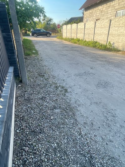 This image captures a dirt road in the village of Мухавецкі сельскі Савет, Belarus, on a clear day with abundant sunlight. On the left side of the road, a dark-colored SUV is parked near a green tree and a red stop sign. Adjacent to the road is a grey concrete fence with a decorative pattern, running parallel to the street. A residential building constructed with light grey bricks is visible in the background, partially obscured by the fence. The road itself is covered in gravel and dirt, with patches of green grass growing along the edges. A dark metal fence is also visible on the left side of the image, positioned in front of a grey pole.
