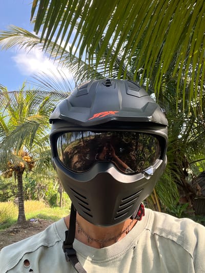 A close-up, eye-level, outdoor selfie shows a person wearing a matte black full-face motorcycle helmet with a dark tinted visor. The helmet has an orange "JX" logo on the side and ventilation grates. The visor reflects a wooded area and the sky with some clouds. The person is wearing a light olive-green t-shirt with distressed detailing around the collar and holes on the shoulder. A thin silver chain with small spike-shaped pendants is visible around their neck. The background features lush green palm fronds in the foreground, with a tropical landscape behind, including palm trees with coconuts and a rice field under a clear blue sky. This scene is located in the city of Kerambitan, Indonesia.