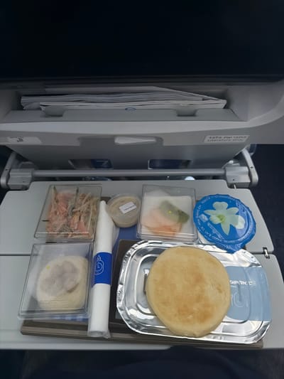 The image shows a tray of food on an airplane. The tray includes a round flatbread, a container of water, a container of tahini sauce, and several other small containers with food items in them. There is also a rolled napkin. The tray is attached to the back of an airplane seat. A label on the seat is written in Hebrew and English.