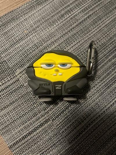 A yellow, Spongebob-shaped keychain holder is positioned on a grey, textured surface. The holder has a black jacket design with the word "STYLE" and the letter "N" visible. A silver carabiner is attached to the top left of the holder. The background is a close-up of a woven mat with wooden flooring visible in the top left corner. The image was taken in Ząbki, Poland.