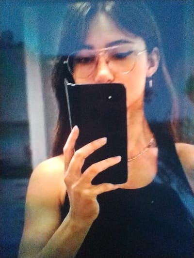 The image shows a woman taking a selfie in what appears to be an indoor setting. The woman is holding a black smartphone in her right hand, angled to capture her reflection. She is wearing glasses and a delicate necklace. Her left hand is visible, with its fingers gracefully spread. The background is dark and out of focus, with some blueish hues suggesting ambient lighting or possibly a screen. The image is laterally inverted, consistent with a front-facing camera selfie. There is no visible text. The overall mood is casual, as indicated by the selfie activity. Without further cues, the exact time of day, weather, or specific location within Brashlen, Bulgaria cannot be determined, other than it is an indoor setting.