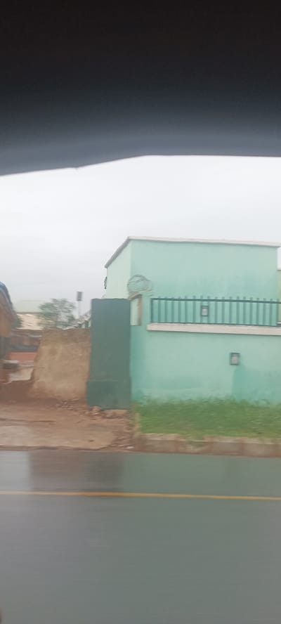 The image depicts a section of a street in what appears to be Umuahia, Nigeria, on an overcast day. The road is wet, and a yellow line marks the lane. On the right, there is a building with a light blue facade and a dark green barred fence. There's grass growing along the base of the fence. Adjacent to the building, there's a section of exposed earth or partially demolished wall, appearing to be made of a reddish-brown material. In the background, other structures are visible, but they are out of focus due to the camera's depth of field and distance. The very top of the frame is obscured by a dark object, likely part of the interior of the vehicle from which the photo was taken. There are no visible people in the shot.