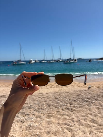 A person's hand holds up sunglasses. The background shows a beach with tan sand and turquoise ocean water. Several sailboats are anchored in the distance, their masts rising into a clear blue sky. The scene is bright and sunny. The sunglasses have brown lenses and a metal frame.