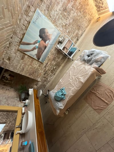 This image depicts an unoccupied treatment room within a spa or wellness facility, located in Kliczków, Poland. The room features exposed reddish-brown brick walls with visible mortar lines, and a light-colored tiled floor composed of rectangular stone slabs.
A beige-upholstered treatment table, covered with a transparent plastic sheet, is centrally positioned. A crumpled light blue towel rests near the head of the table, and a folded light beige towel with an embossed pattern lies on the floor adjacent to the table's right side. A silver-colored rolling swivel stool with a white circular seat is situated at the foot of the treatment table.
Mounted diagonally on the brick wall is a large framed poster advertising "THALGO SPA." The poster illustrates a female subject from the back, receiving a cupping therapy treatment from two sets of hands, which are applying blue suction cups to her upper back.
Adjacent to the poster on the right, a white, three-tiered utility cart with wheels holds several folded beige and blue towels, along with various bottles and containers on its top shelf.
In the lower-left section of the image, a white countertop with an integrated oval basin and a silver faucet is visible. A wooden cabinet is situated beneath the counter. Items on the countertop include a small green potted plant, various bottles, and a folded blue towel. A gold-framed mirror is partially visible on the far left, reflecting a portion of the room. The ceiling exhibits an arched brick structure in places, and several recessed lighting fixtures illuminate the space. A dark, circular fixture, possibly a vent or another light, is mounted high on the right wall/ceiling. The room appears prepared for use, with no people present.