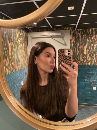 A woman is taking a selfie in a bathroom mirror. She is holding a phone with a leopard print case. The bathroom has a mix of black and white checkered tile, wallpaper with a gold and green leaf pattern, and blue tile. There is a closed white door visible in the mirror. The location is likely in Uppsala, Sweden, as the user mentioned the city in the request.