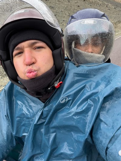 The image shows a close-up view of two individuals on a motorcycle in the city of Sumdo, India. Both individuals are wearing helmets and raincoats, indicating wet weather conditions, likely rain, as evidenced by the water droplets on the helmets' visors and the damp appearance of the raincoats. The person in the foreground, wearing a maroon helmet and a dark neck gaiter, is puckering their lips and looking towards the camera. The second person, behind the first and wearing a dark blue helmet, is partially obscured by the first person's helmet and is wearing a light-colored face mask. The background suggests a mountainous or hilly terrain, with a road visible. The overall scene depicts a moment captured during a motorcycle ride in inclement weather in Sumdo.